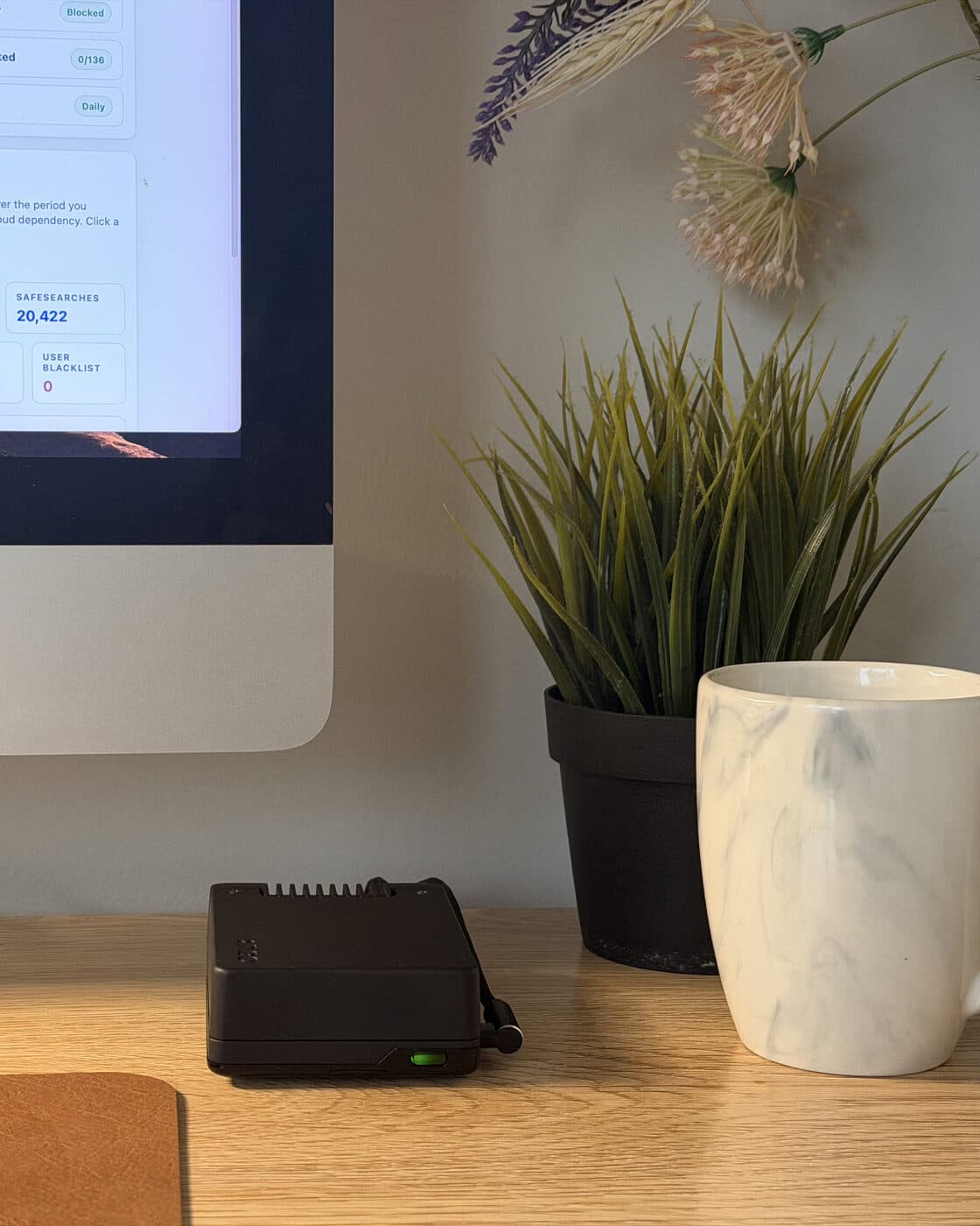 Shield Core on an oak desk beside a plant and a mug, green status light on, with the Shield dashboard open on the iMac behind it.