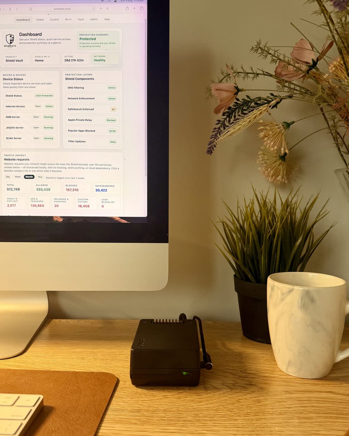 Shield Vault on an oak desk, green status light on, with its dashboard open on the iMac behind it showing 28 days uptime and every protection layer active.