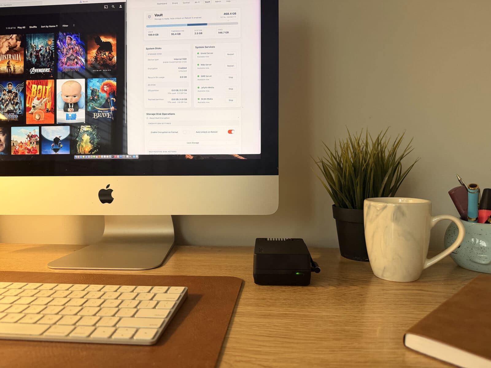 An iMac split between the household's own film library and Shield Vault's storage page — 468.4 GB total, encryption enabled — with the Vault itself on the desk beside the keyboard, green status light on.